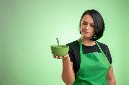 Female cook with green apron and black t-shirt, she looks at the bowl she is holding in her hand isolated on green backgroundの写真素材