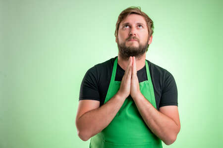 Supermarket employee with green apron and black t-shirt, praying isolated on green backgroundの写真素材