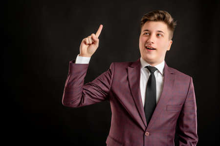 Law student with blond hair dressed in burgundy jacket, white shirt and black tie his finger up has come up with an idea posing on isolated black background with copy space advertising areaの写真素材