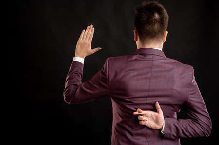 Law student with blond hair dressed in burgundy jacket, white shirt and black tie showing fake oath cross finger from behind posing on isolated black background with copy space advertising areaの写真素材