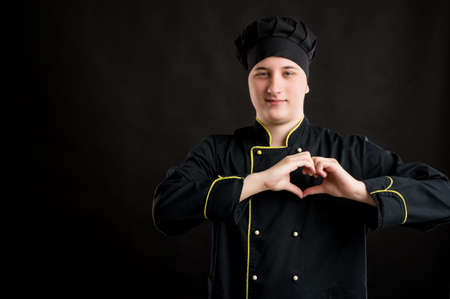 Portrait of young male dressed in a black chef suit showing heart sign with hands posing on a black isolated background with copy space advertising areaの写真素材