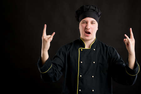 Portrait of young male dressed in a black chef suit doing rock symbol with hands up posing on a black isolated background with copy space advertising areaの写真素材