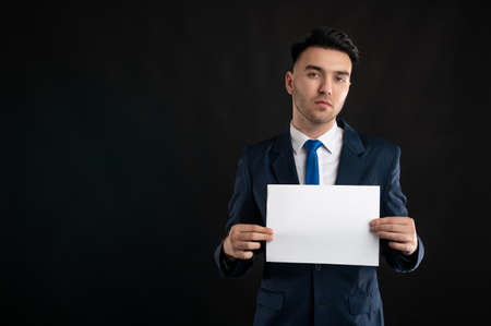 Portrait of business man wearing blue business suit and tie holding white paper isolated on black background with copy space advertising areaの写真素材