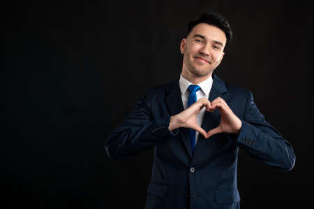 Portrait of business man wearing blue business suit and tie making heart gesture isolated on black background with copy space advertising areaの写真素材