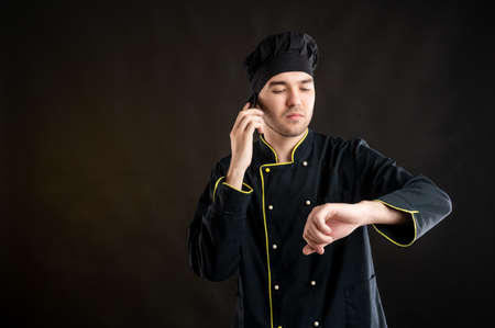 Portrait of young male dressed in a black chef suit showing it's time gesture and looking his watch posing on a brown background with copy space advertising areaの写真素材