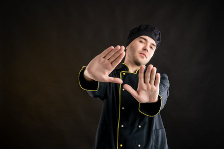Portrait of young male dressed in a black chef suit showing stop sign with boath hands posing on a...