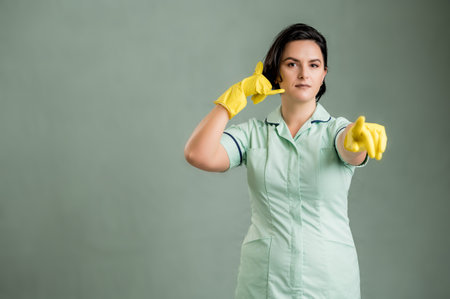 Young cleaning woman wearing a green shirt and yellow gloves, showing call me gesture, pointing her...