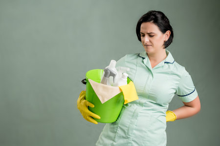 Young cleaning woman wearing a green shirt and yellow gloves, holding cleaning tools and products in...