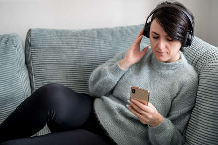 A young woman is resting on a sofa with a smartphone in her hand, listening good musicの写真素材