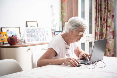 An elder lady with glasses staring at laptop's monitor while clicking with a mouseの写真素材
