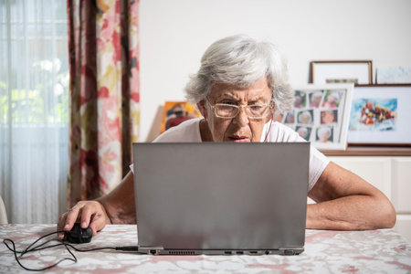 An elder lady with glasses staring at laptop's monitor while clicking with a mouse