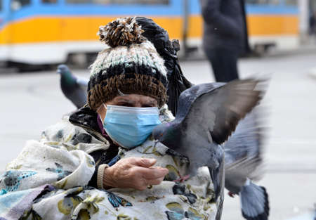 Sofia, Bulgaria - Nov 10 2020: A homeless woman in protective mask is feeding the pigeons during the corona virus outbreakのeditorial素材