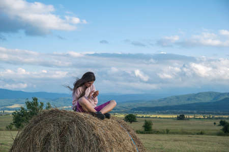 A girl sitting on a stack of hay against cloudy skyの写真素材
