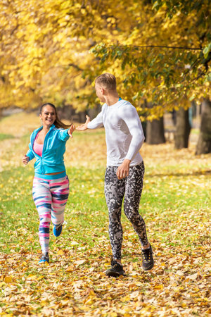 Beautiful young couple running together in the park. Man helps woman giving his hand. Autumn environment.の写真素材