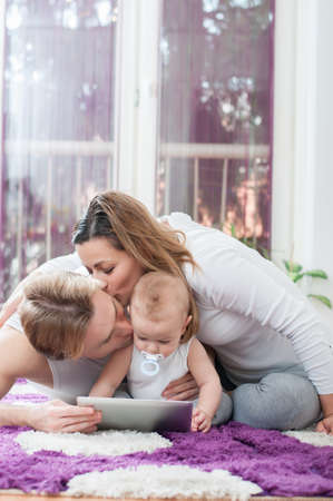 Happy parents laying down on the floor and playing on tablet with their baby boy. Mother kissing a father.の写真素材