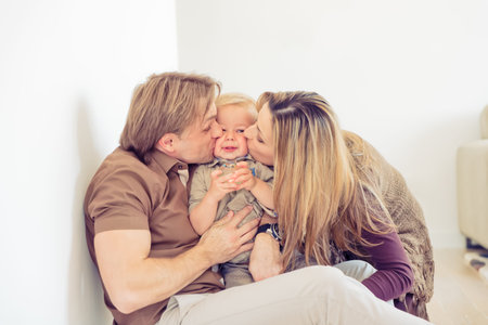 Happy family sitting on floor with their little baby. Family spending time at home with their son. Parents kissing their child.の写真素材