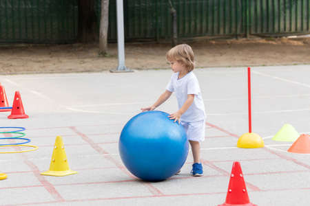 Beautiful little boy rolling the big blue ball on sports polygonの写真素材