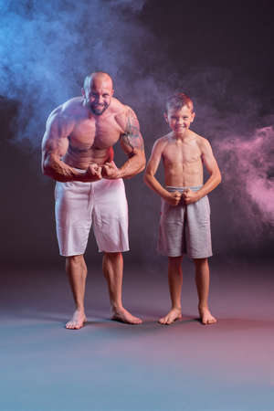 Powerful muscular man shirtless posing with his son in the studio with red and blue smokeの写真素材