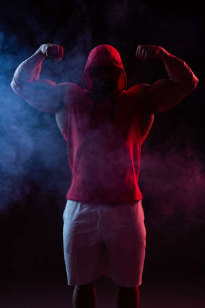 Powerful muscular man with a hood posing in the red sweatshirt. Studio shoot with red and blue smokeの写真素材