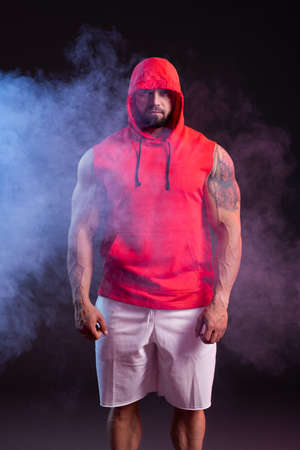Powerful muscular man with a hood posing in the red sweatshirt. Studio shoot with red and blue smokeの写真素材