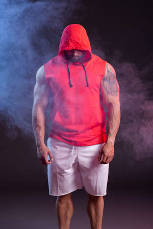 Powerful muscular man with a hood posing in the red sweatshirt. Studio shoot with red and blue smokeの写真素材