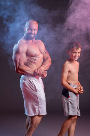 Powerful muscular man shirtless posing with his son in the studio with red and blue smokeの写真素材