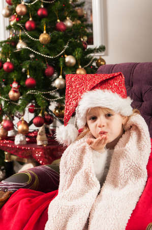 Beautiful little girl with santa claus hat wrapped into red blanket, sitting beside christmass tree and sending kissesの写真素材