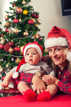 Mother and her beautiful baby boy posing beside christmass tree with santa claus hatsの写真素材