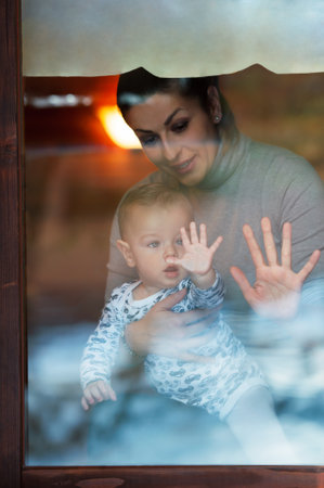 Happy mother and her baby boy looking throw the window and playing in warm home on winter day. Christmas time.の写真素材