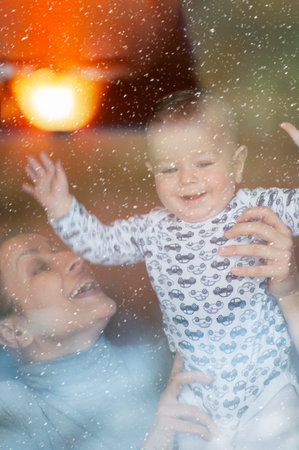 Happy mother and her baby boy looking throw the window and playing in warm home on winter day. Christmas time.の写真素材