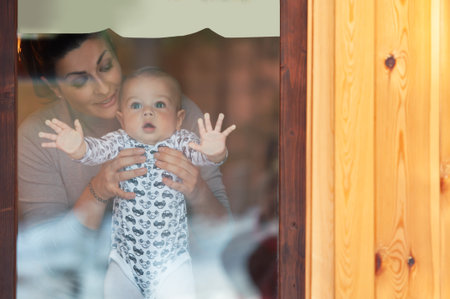 Happy mother and her baby boy looking throw the window and playing in warm home on winter day. Christmas time.の写真素材