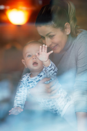 Happy mother and her baby boy looking throw the window and playing in warm home on winter day. Christmas time.の写真素材