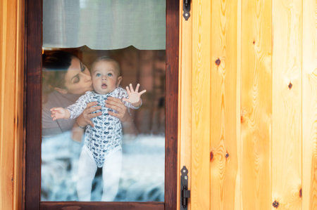 Happy mother and her baby boy looking throw the window and playing in warm home on winter day. Christmas time.の写真素材