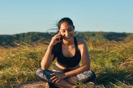 A happy young woman sitting on the grass and talking on the phone after workout
