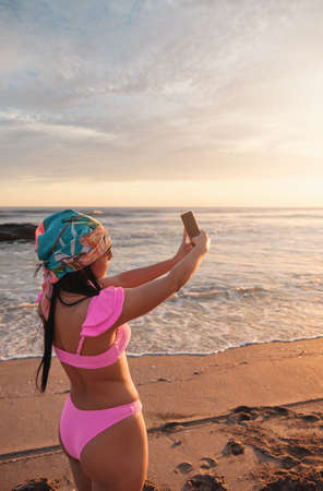 A gorgeous latina woman wearing a pink bikini and a bandana taking selfie with smartphone in a beautiful sunset on the sea shore.の写真素材