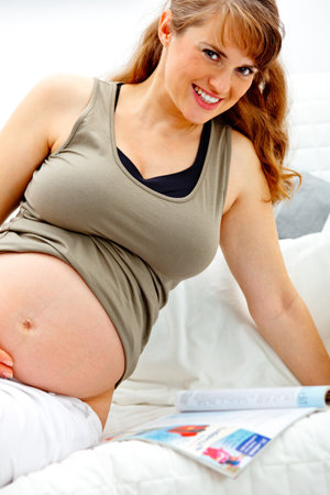 Smiling beautiful pregnant woman sitting on sofa at home with magazine.