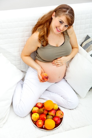 Smiling  beautiful pregnant female sitting  on couch and  holding fruit in hand
の写真素材