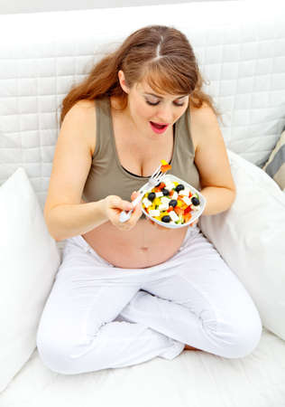 Smiling beautiful pregnant woman sitting on sofa at home and eating fresh vegetable saladの写真素材