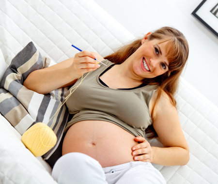Smiling beautiful pregnant  female sitting on sofa and knitting for her baby.の写真素材