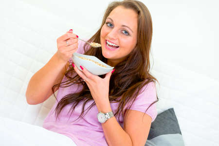 Smiling beautiful woman sitting on sofa  at home and eating muesliの写真素材