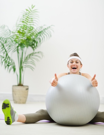 Smiling woman sitting behind fitness ball and showing thumbs upの写真素材