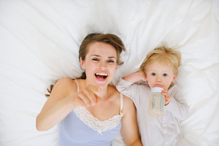 Mother laying with baby in bed and pointing in camera.