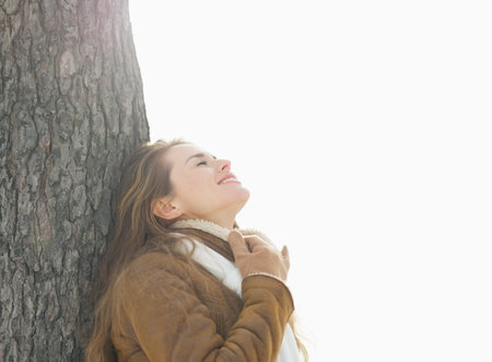 Happy young woman leaning against tree in winter parkの写真素材