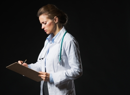 Serious doctor woman writing in clipboard isolated on blackの写真素材