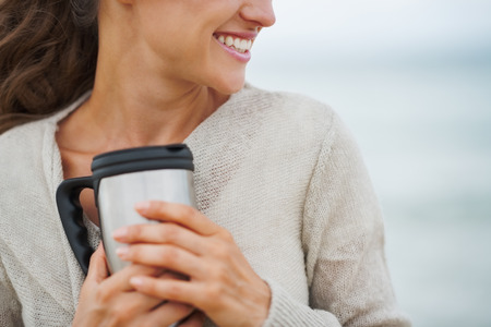 Closeup on happy young woman in sweater on beach with cup of hot beverageの写真素材