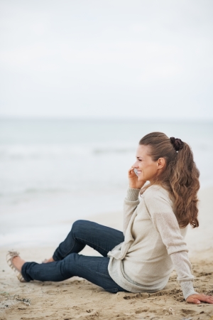 Happy young woman in sweater sitting on lonely beach and talking cell phoneの写真素材