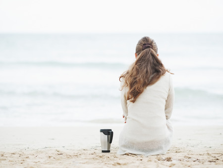 Cup of hot beverage near young woman in sweater sitting on lonely beach   rear viewの写真素材