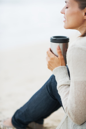 Young woman in sweater sitting with cup of hot beverage and relaxing on lonely beachの写真素材
