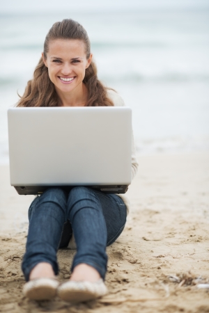 Smiling young woman in sweater sitting on lonely beach with laptopの写真素材