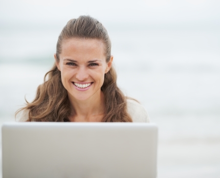 Portrait of smiling young woman in sweater sitting on beach with laptopの写真素材